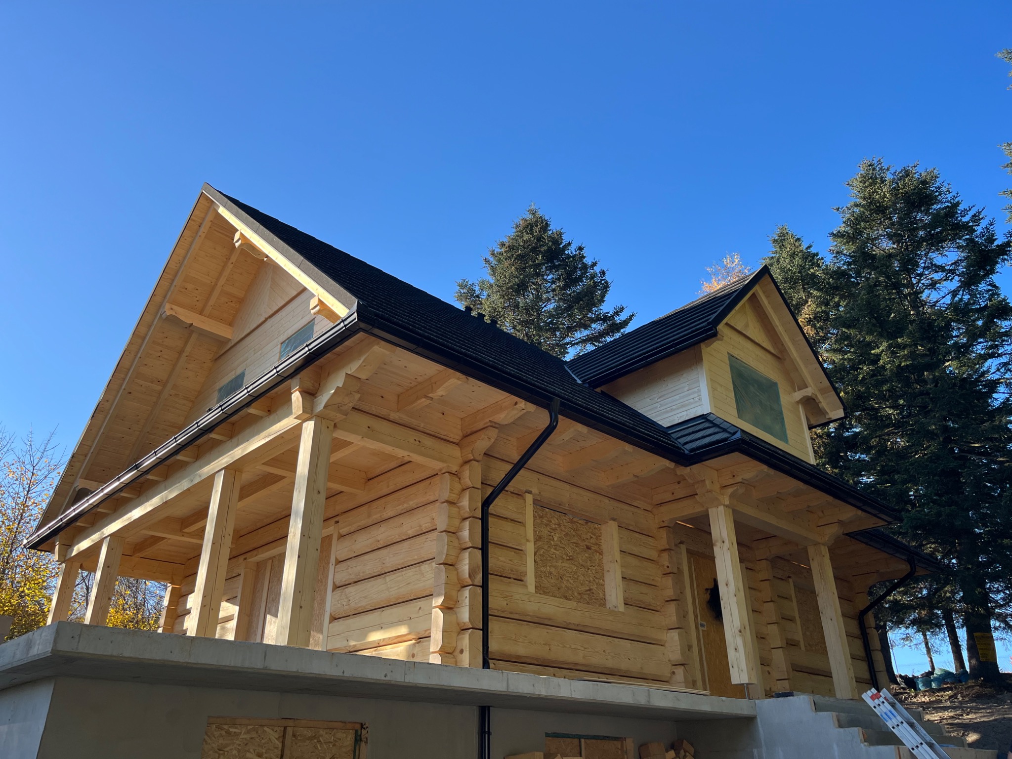 Surowy stan drewnianego domu z bali z czarnym dachem, widoczne deski zakrywające okna i szara podmurówka, w tle drzewa iglaste i błękitne niebo.