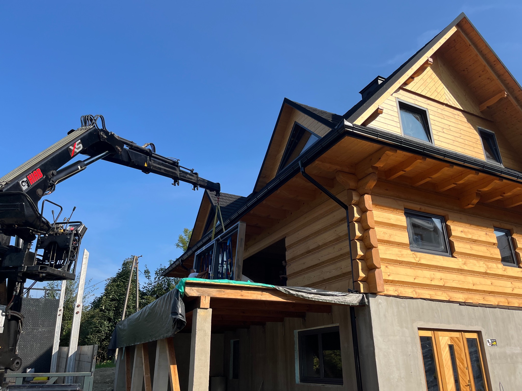 Montaż okna w nowym domu z bali za pomocą dźwigu HIAB XS, widoczna konstrukcja z drewnianych bali i betonowy fundament