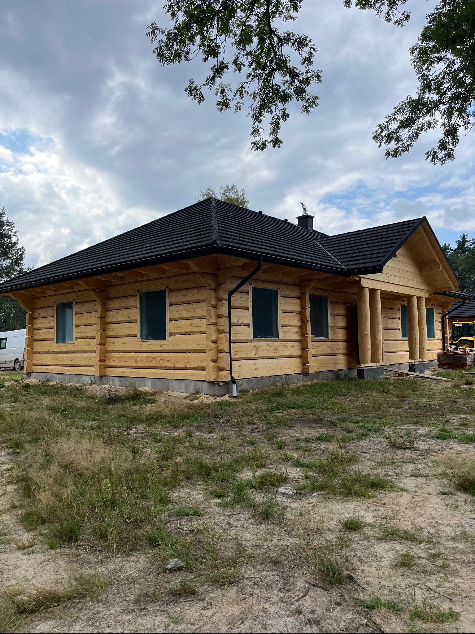 Surowy, jasny drewniany dom z bali z czarnym dachem, jeszcze bez wykończenia elewacji, z widocznym transportem i koparką w tle.