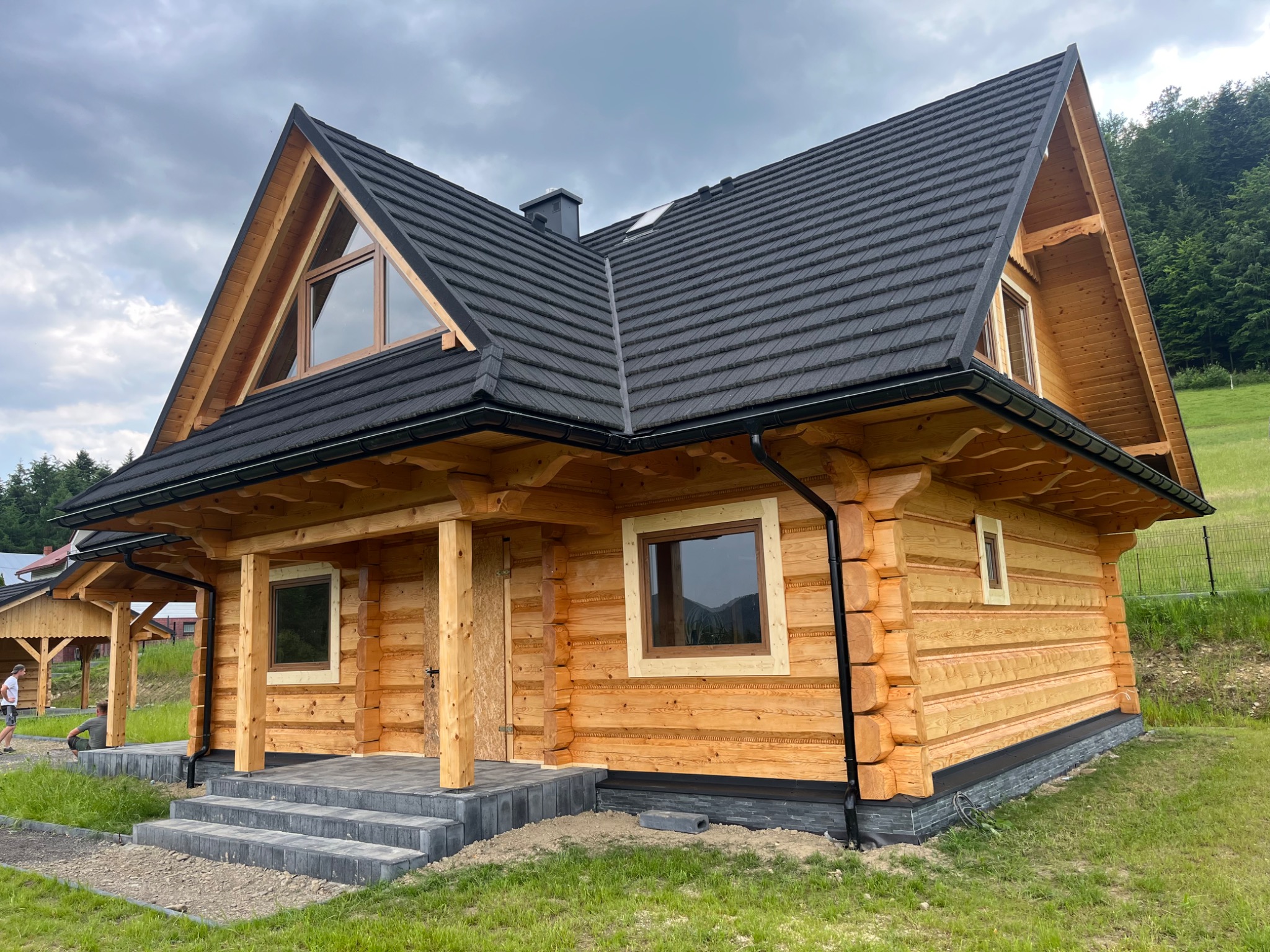 Nowoczesny dom z bali z czarnym dachem i drewnianymi detalami, otoczony zielenią. Widoczne schody z szarej kostki i fragment balustrady. Dachówka w ciemnym kolorze.