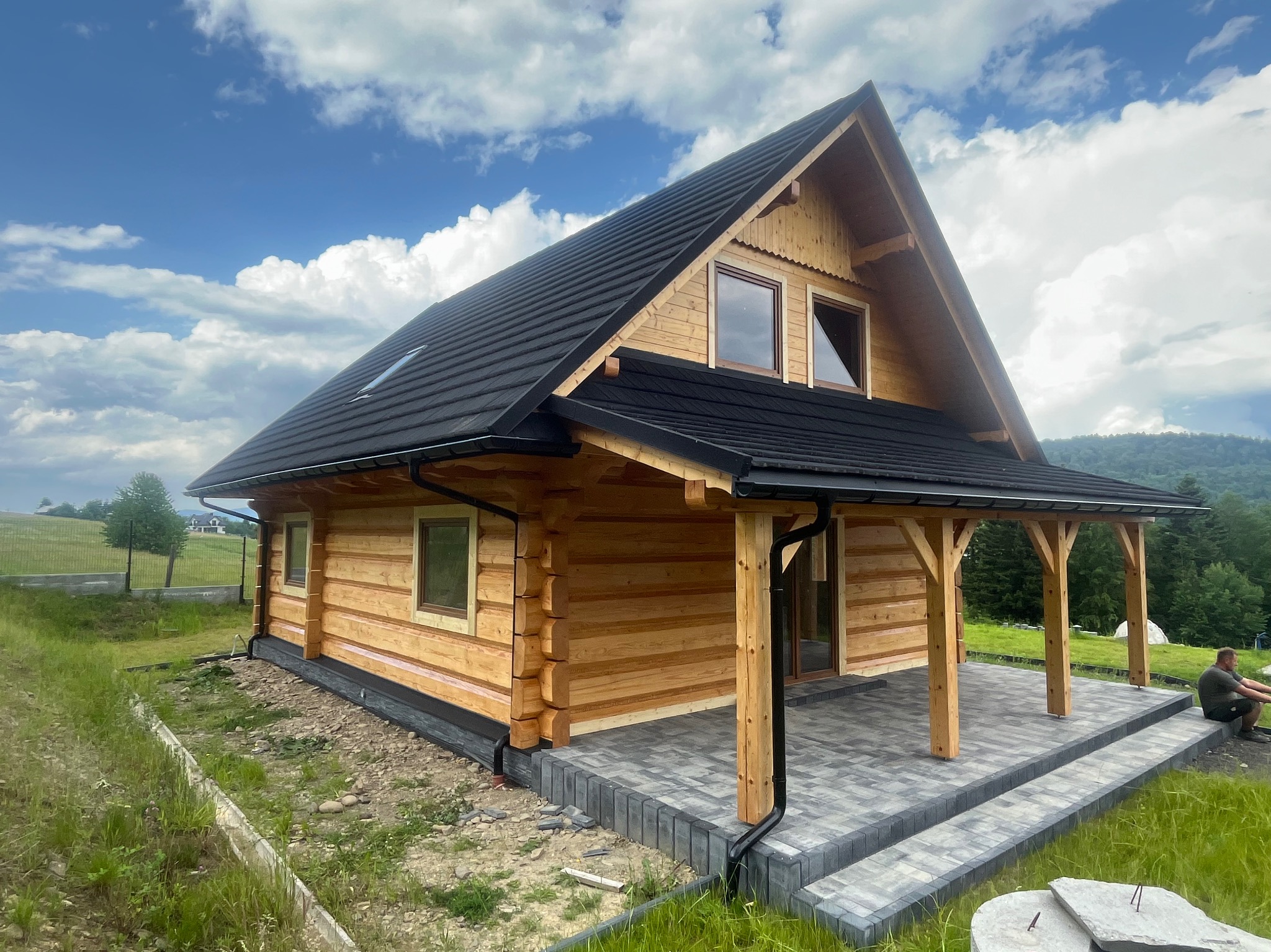 Nowoczesny dom z bali z czarnym dachem i zadaszonym tarasem wyłożonym szarą kostką, na tle błękitnego nieba z chmurami i zielonych wzgórz.