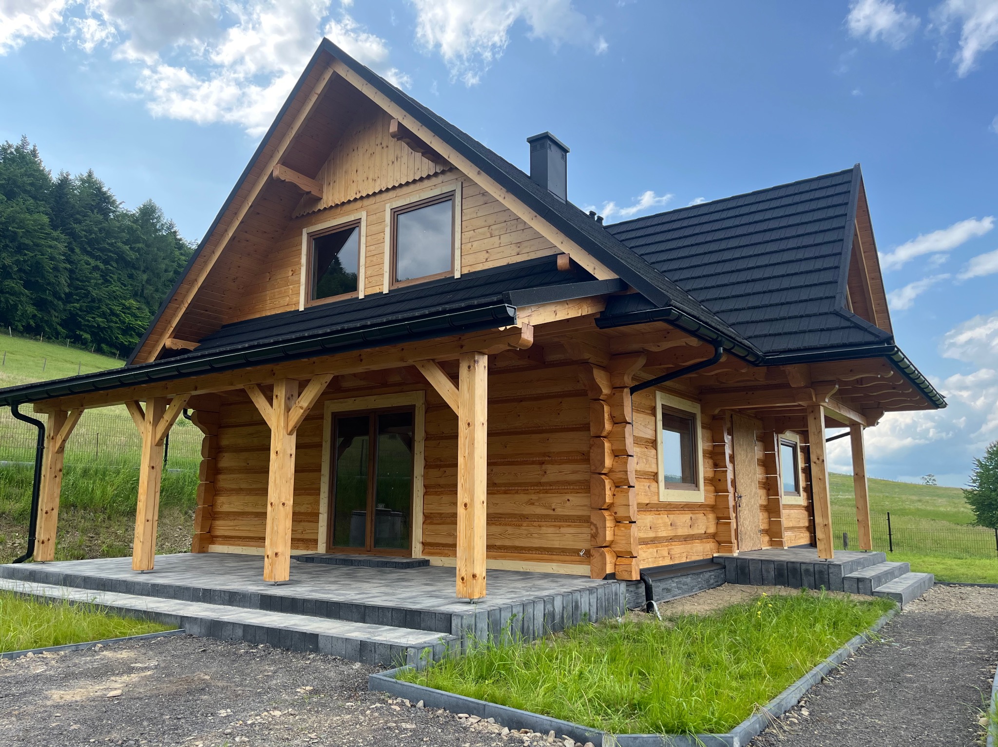 Nowoczesny dom z bali z ciemną dachówką i jasnym drewnem, zadaszony taras z kamienną posadzką, zielony trawnik. Ujęcie w słoneczny dzień.