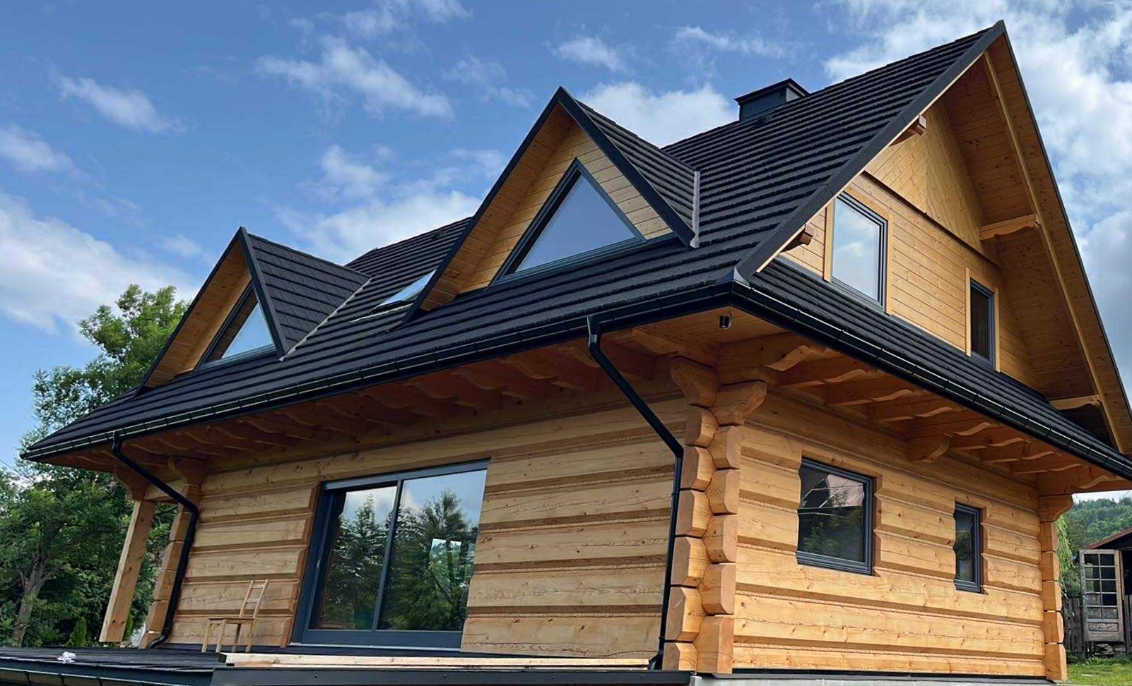 Nowoczesny dom z bali z czarnym dachem i oknami. Widoczny fragment elewacji z charakterystycznymi połączeniami bali i trójkątne okno na poddaszu. W tle błękitne niebo.