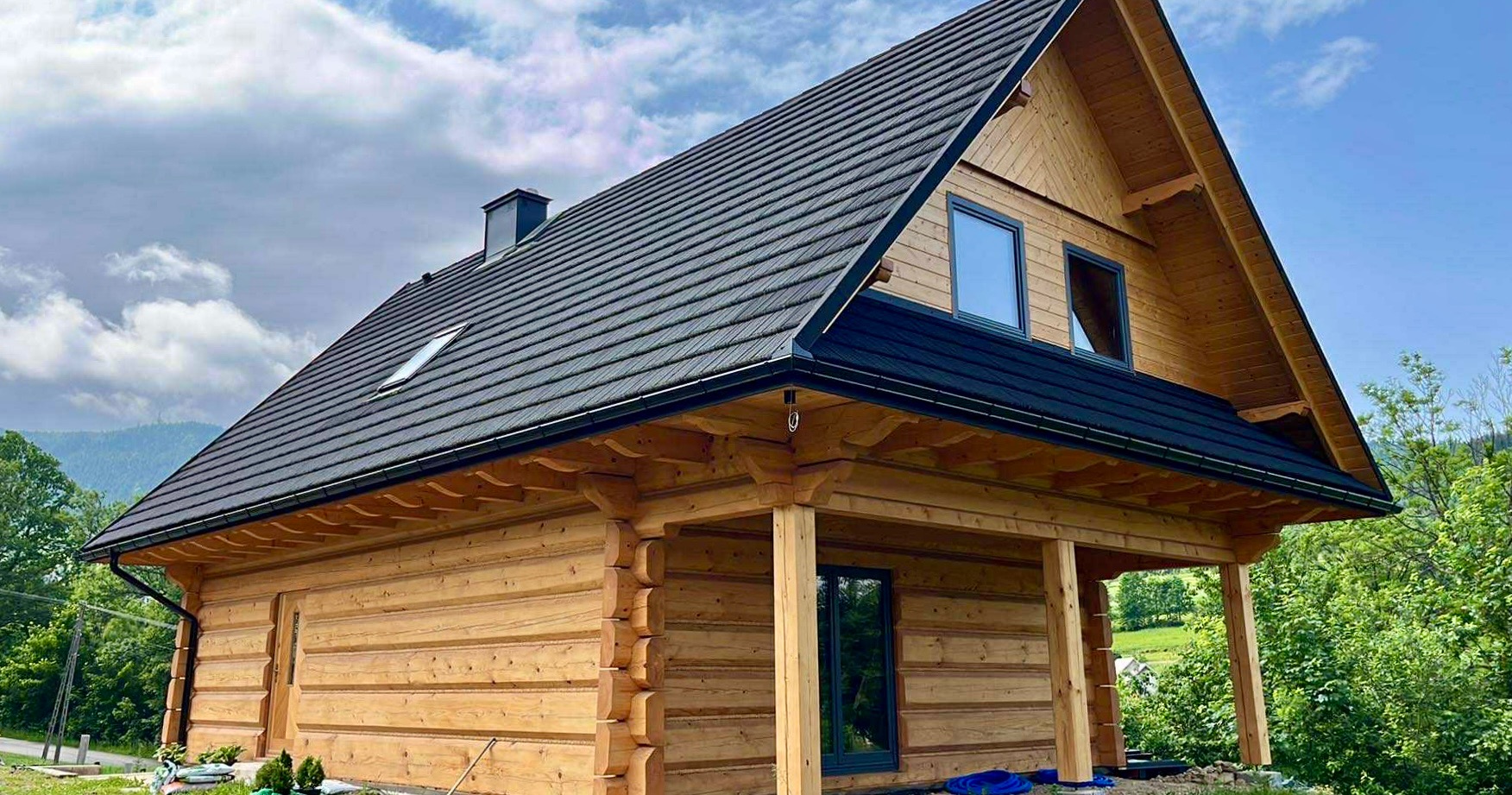 Ukończony, parterowy dom z bali z ciemnym dachem, widoczny z zewnątrz, z elementami wykończeniowymi i fragmentem zielonego otoczenia.