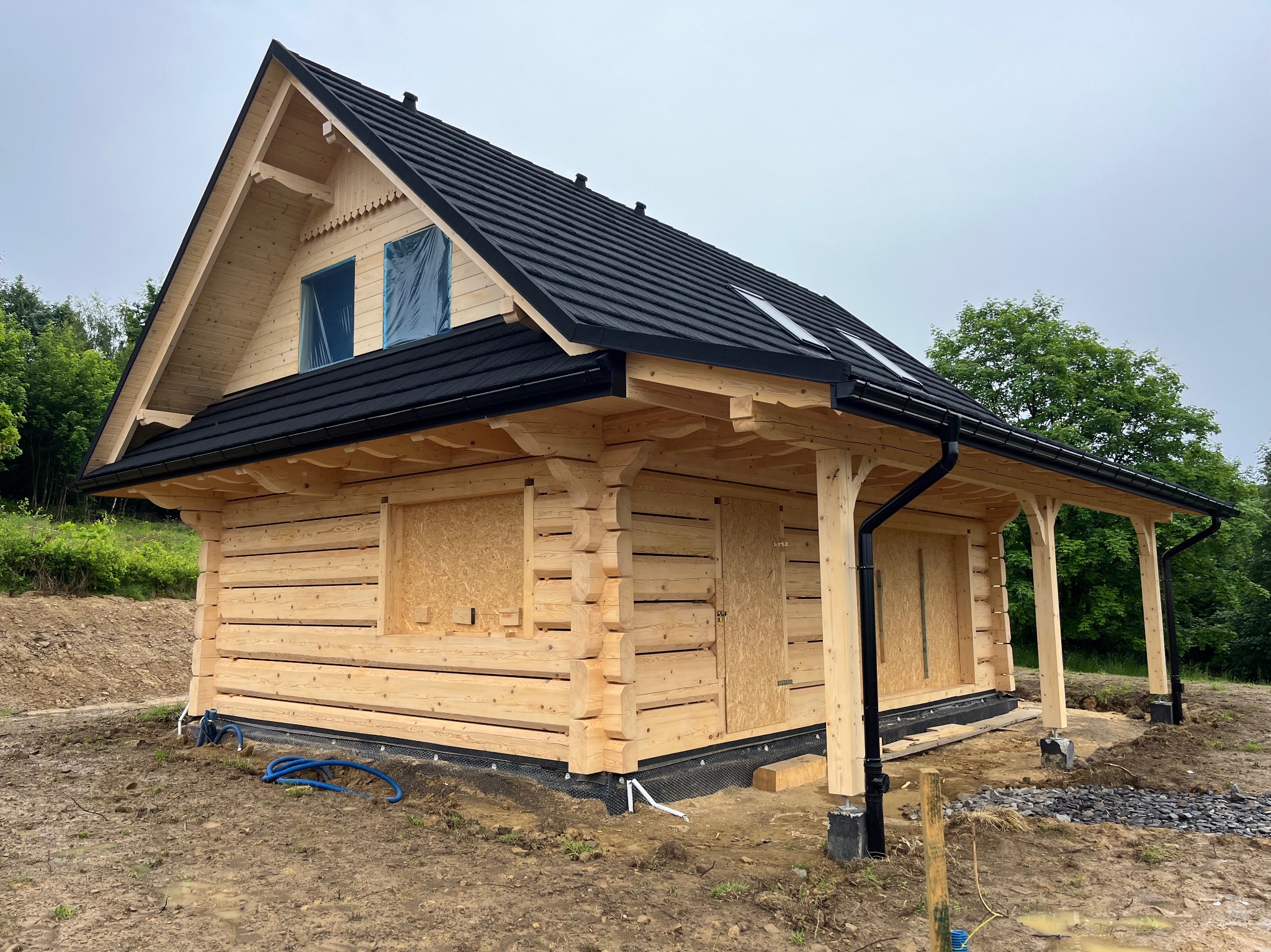 Surowy stan domu z bali z czarnym dachem, okna zabezpieczone folią, widoczne fundamenty i teren budowy. Drewniana konstrukcja z bali, świeże drewno, surowy krajobraz.