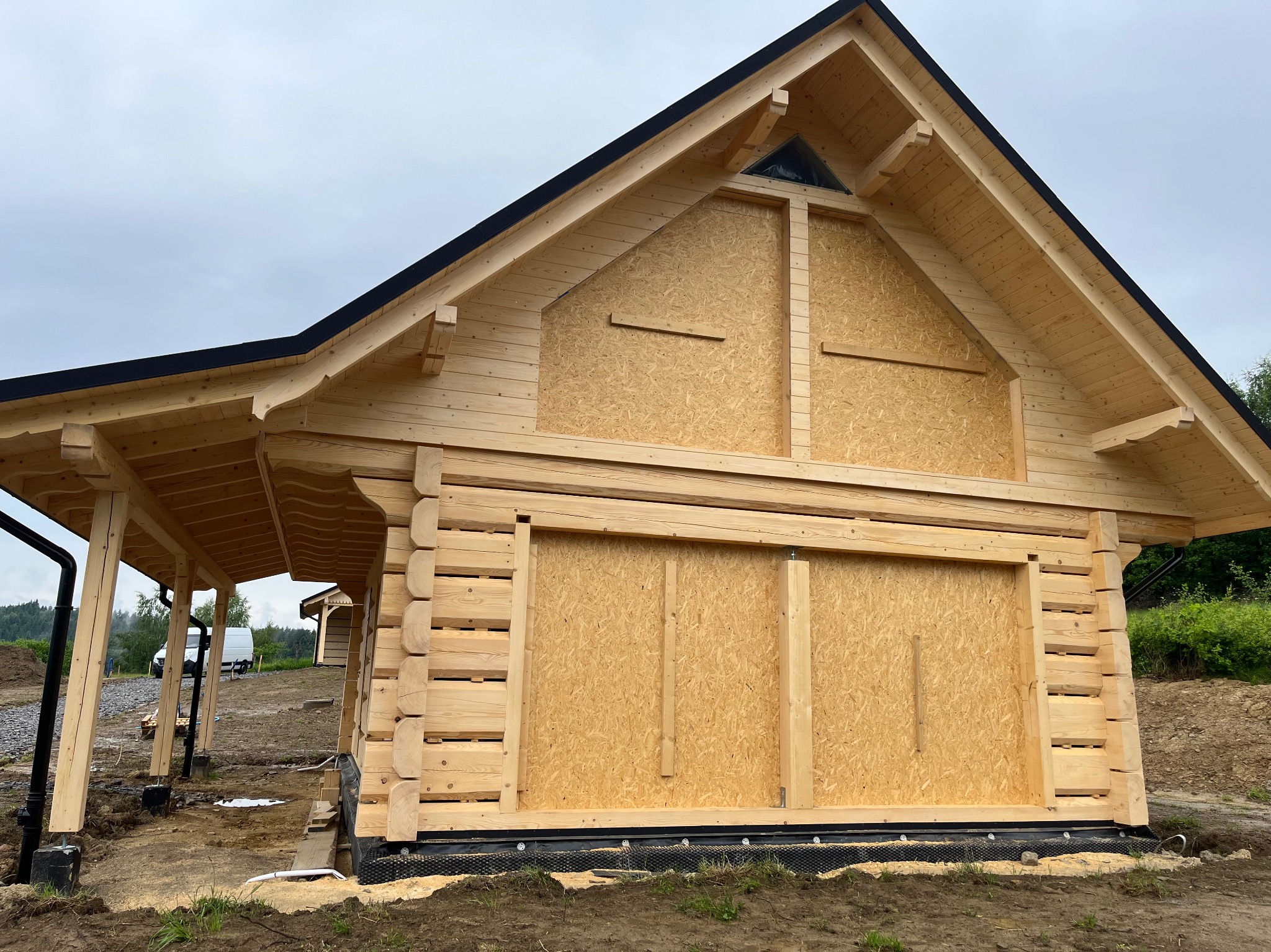 Surowy stan domu z bali w trakcie budowy, z deskami OSB zamiast okien, widok z zewnątrz na tle pochmurnego nieba. Konstrukcja z jasnego drewna.