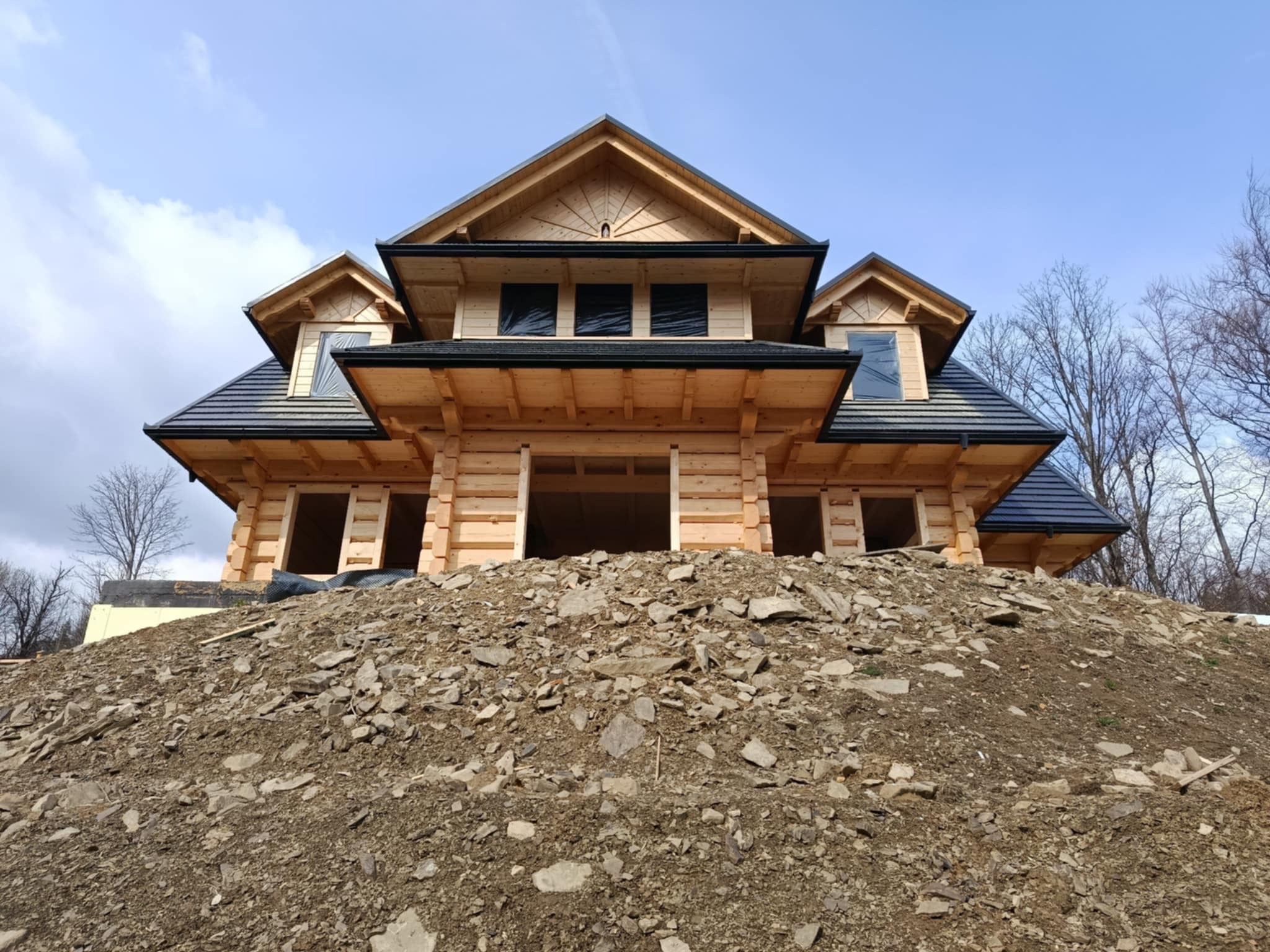 Surowy stan budowy domu z bali, z widoczną konstrukcją drewnianą, czarnym dachem i przygotowanym terenem pod budowę na tle błękitnego nieba.