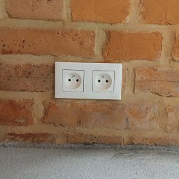 Kompleksowe Usługi Elektryczne Elektro-Lib Jakub Liberacki - Podwójne gniazdko elektryczne w białej ramce, zamontowane na ścianie z czerwonej cegły z widocznymi spoinami, poniżej szara, surowa ściana.