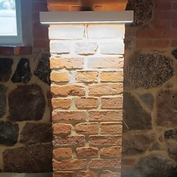 Kompleksowe Usługi Elektryczne Elektro-Lib Jakub Liberacki - Oświetlony fragment ceglanej kolumny z podświetleniem LED, przylegającej do ściany z kamienia i cegły, widoczne okno w tle.