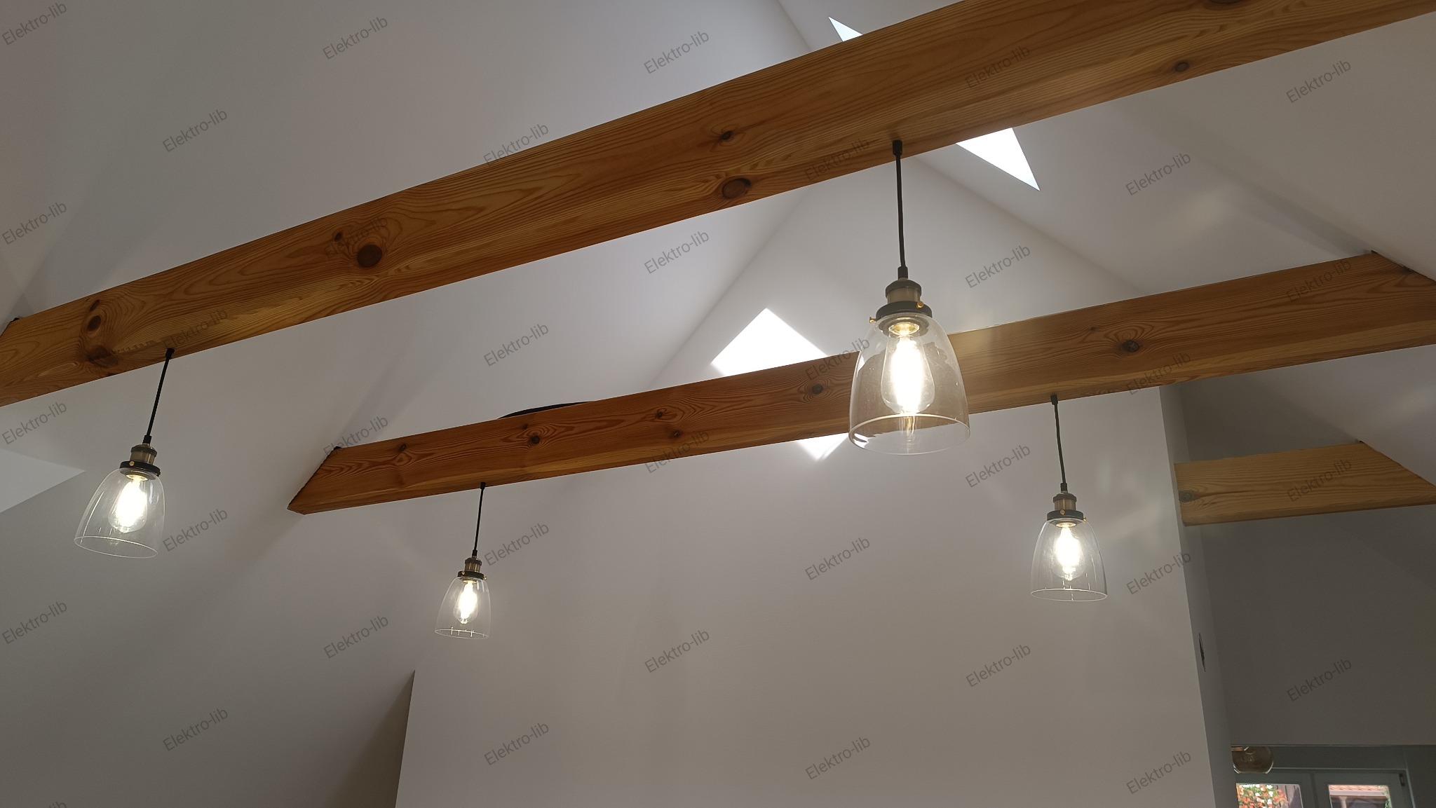 Cztery szklane lampy wiszące z żarówkami Edison zamontowane na drewnianych belkach stropowych w jasnym pomieszczeniu z oknem dachowym.