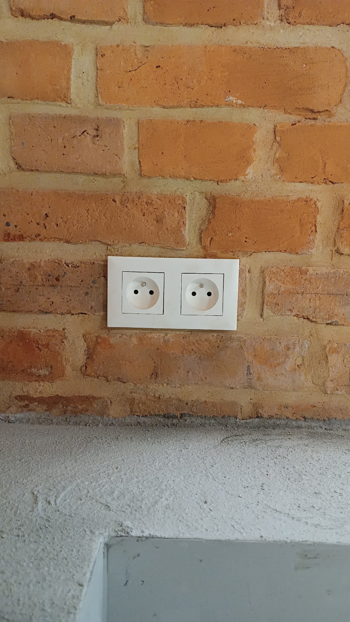 Podwójne gniazdko elektryczne w białej ramce, zamontowane na ścianie z czerwonej cegły z widocznymi spoinami, poniżej szara, surowa ściana.