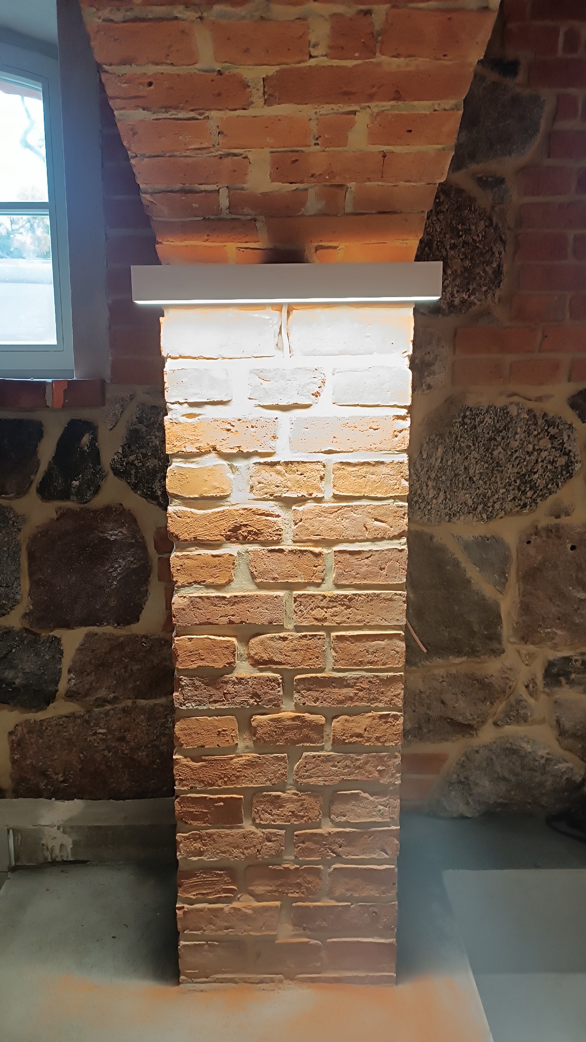 Oświetlony fragment ceglanej kolumny z podświetleniem LED, przylegającej do ściany z kamienia i cegły, widoczne okno w tle.