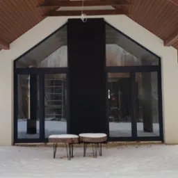 Nowoczesny dom z dużymi, przesuwnymi drzwiami tarasowymi w czarnych ramach, widok z zewnątrz w zimowej scenerii, dwa małe stoliki z drewnianymi blatami na śniegu.