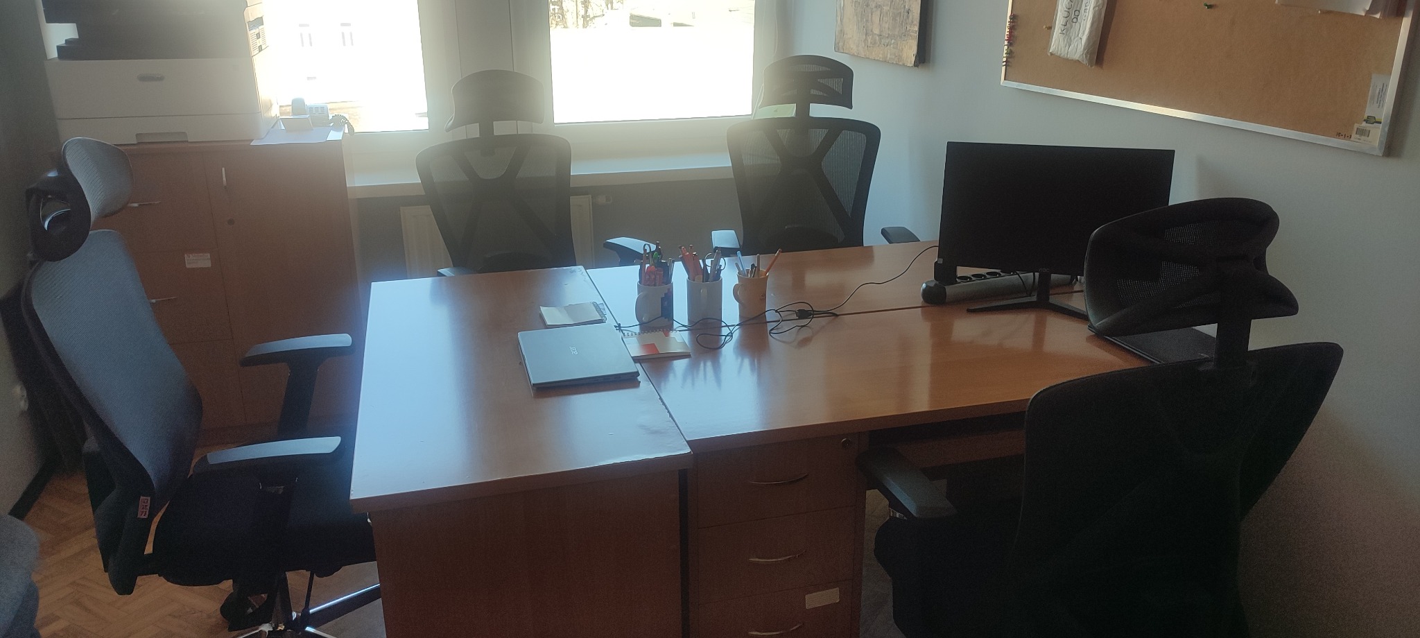 Biuro z trzema krzesłami z siatkowym oparciem, biurkiem z drewnianym blatem, komputerem, laptopem i organizerami na długopisy, w tle szafka i tablica korkowa.