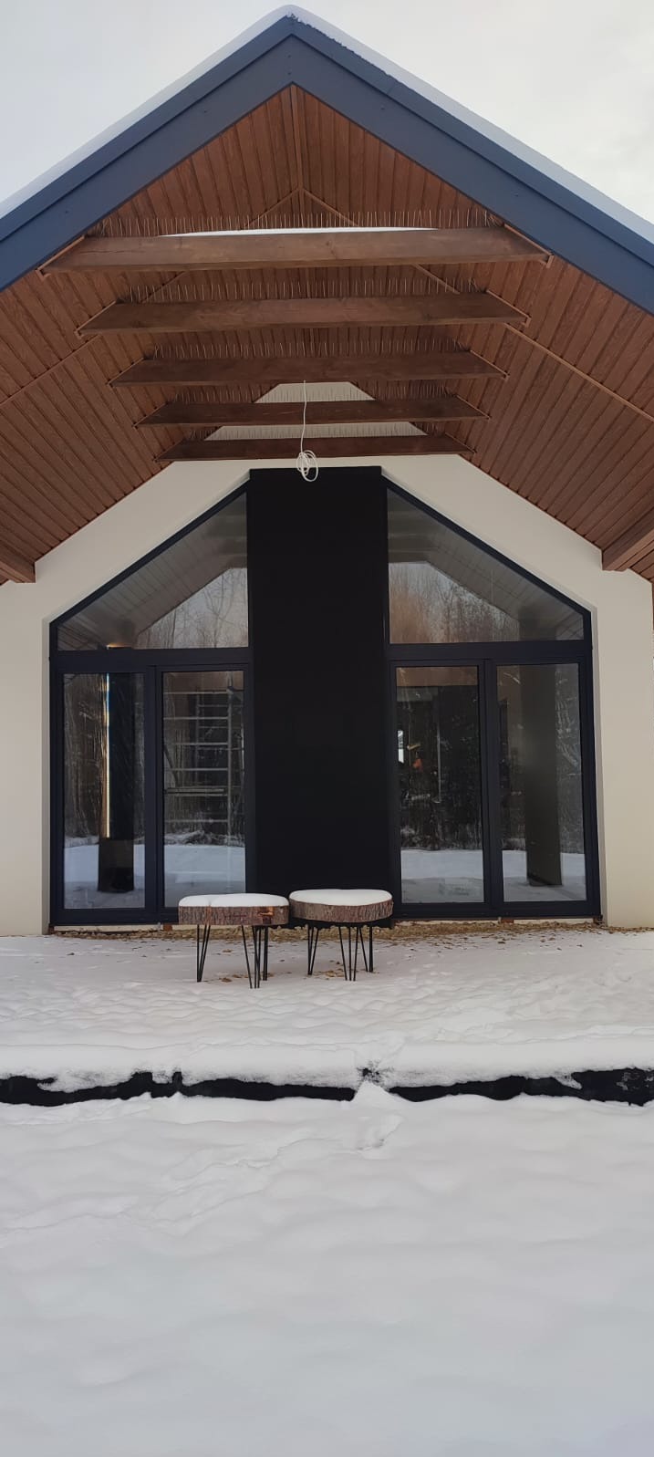 Nowoczesny dom z dużymi, przesuwnymi drzwiami tarasowymi w czarnych ramach, widok z zewnątrz w zimowej scenerii, dwa małe stoliki z drewnianymi blatami na śniegu.