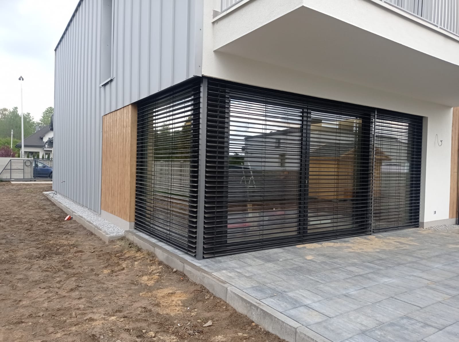 Zewnętrzne, poziome żaluzje fasadowe w kolorze czarnym, zamontowane na nowoczesnym budynku z szarą elewacją i drewnianymi panelami; widoczny fragment kostki brukowej i przygotowany teren.