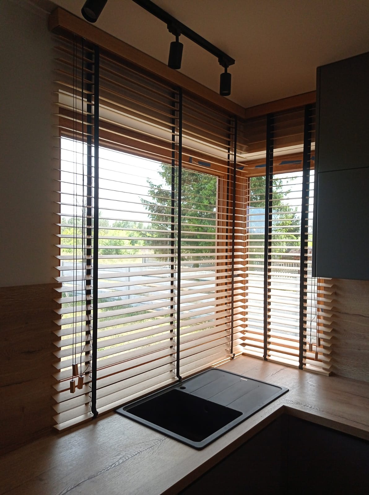 Drewniane żaluzje poziome z czarnymi taśmami w kuchni, częściowo odsłonięte, widok na zieleń za oknem, czarny zlew w blacie.