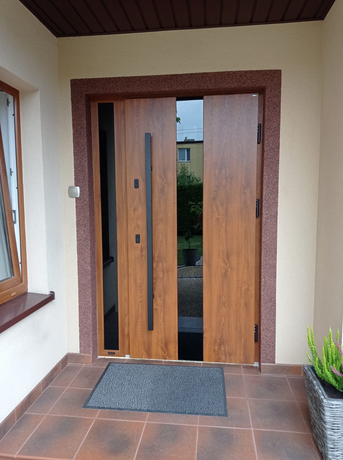 Nowoczesne, dwuskrzydłowe drzwi wejściowe z drewnopodobnym wykończeniem, czarnym, pionowym uchwytem i wąskimi przeszkleniami, umieszczone w elewacji z brązowym obramowaniem i brązowymi płytkami...