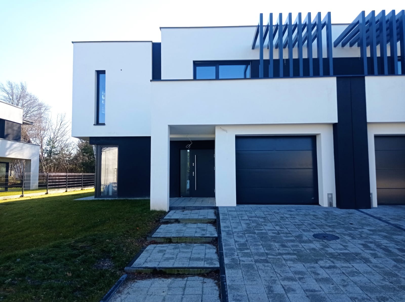 Nowoczesny dom jednorodzinny z czarną stolarką okienną i bramą garażową, widok z zewnątrz na wejście i podjazd z kostki brukowej.