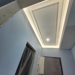 GWARANT - WOWA VOLODYMYR MELNYK - Wykończony sufit z ozdobnymi listwami i oświetleniem w kształcie prostokąta, niebieskie ściany w trakcie zabezpieczania folią malarską, widoczne puszki elektryczne.