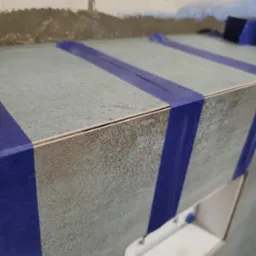 Montaż płytek ceramicznych na zabudowie w łazience, zabezpieczonych niebieską taśmą malarską, widoczne niedoskonałości na ścianie w tle.