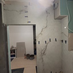 GWARANT - WOWA VOLODYMYR MELNYK - Ściana w trakcie układania płytek imitujących marmur, z użyciem klipsów poziomujących, widoczne płyty kartonowo-gipsowe i przygotowane podłoże w remontowanej łazience.