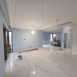 GWARANT - WOWA VOLODYMYR MELNYK - Przestronne wnętrze w trakcie prac wykończeniowych: marmurowe płytki podłogowe, pomalowane na jasny błękit ściany z dekoracyjnymi panelami, surowe przewody zwisające z sufitu, widoczne narzędzia...