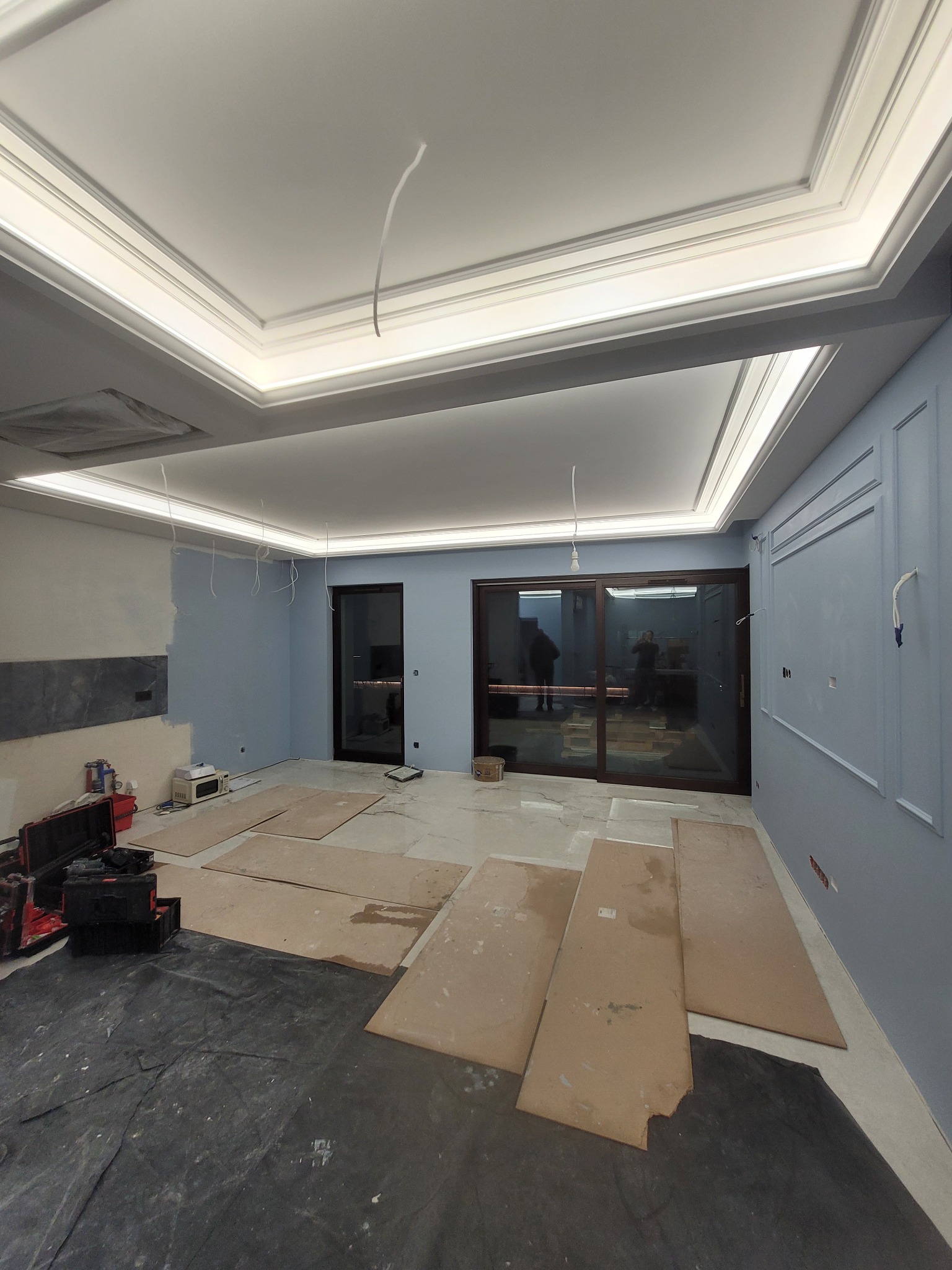 Pokój w trakcie prac wykończeniowych: pomalowane na błękitno ściany z dekoracyjnymi listwami, marmurowe płytki podłogowe częściowo zakryte kartonem, sufit z oświetleniem LED i dekoracyjnymi...