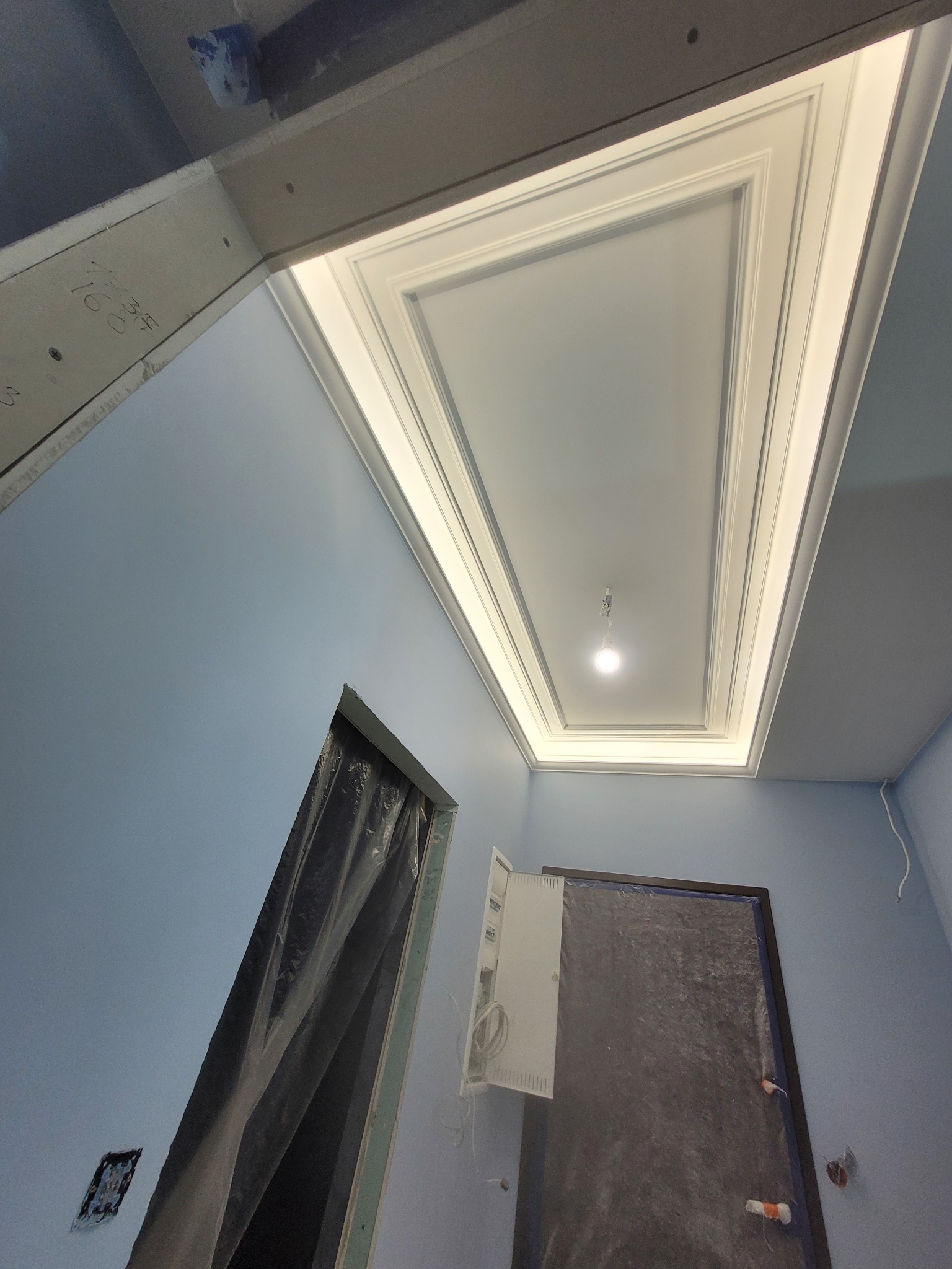 Wykończony sufit z ozdobnymi listwami i oświetleniem w kształcie prostokąta, niebieskie ściany w trakcie zabezpieczania folią malarską, widoczne puszki elektryczne.