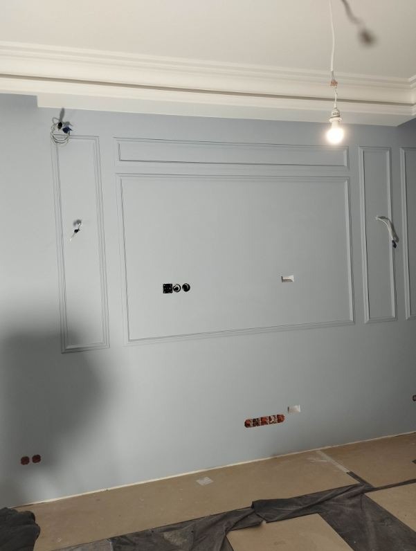 Ściana w trakcie prac wykończeniowych, pomalowana na szaro-niebieski kolor z dekoracyjnymi panelami ściennymi, widoczne gniazdka elektryczne i wystające kable, sufit z ozdobnym gzymsem, podłoga...