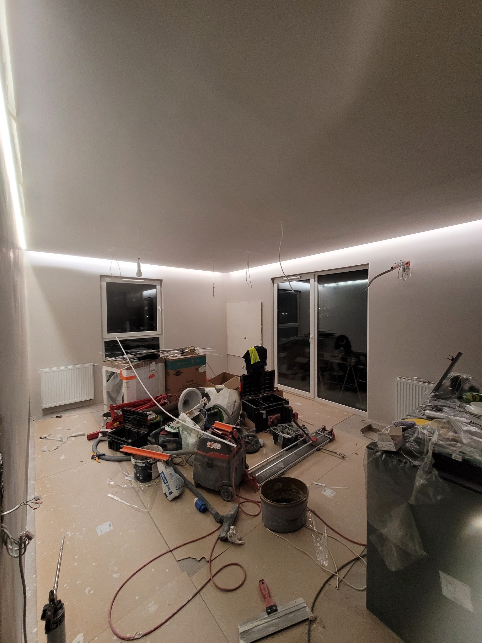 Wnętrze w trakcie prac wykończeniowych: widoczne narzędzia, sprzęt budowlany, sufit z oświetleniem LED, okna i drzwi balkonowe, grzejniki, kable zwisające z sufitu.