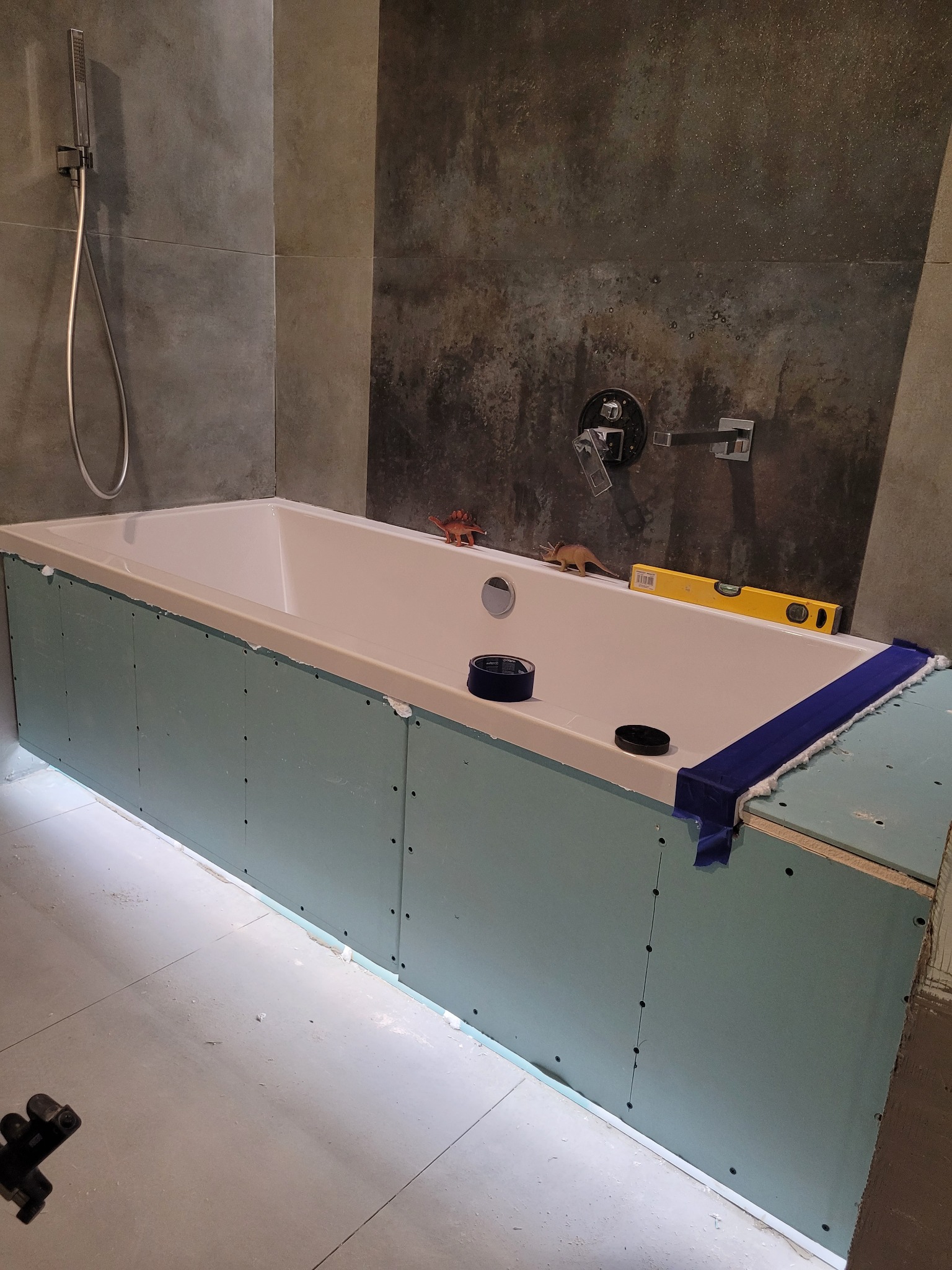 Biała wanna obudowana płytami kartonowo-gipsowymi, widoczne narzędzia: poziomica, taśma malarska, maskownica, zabawki dinozaury na wannie, szare płytki na ścianach.