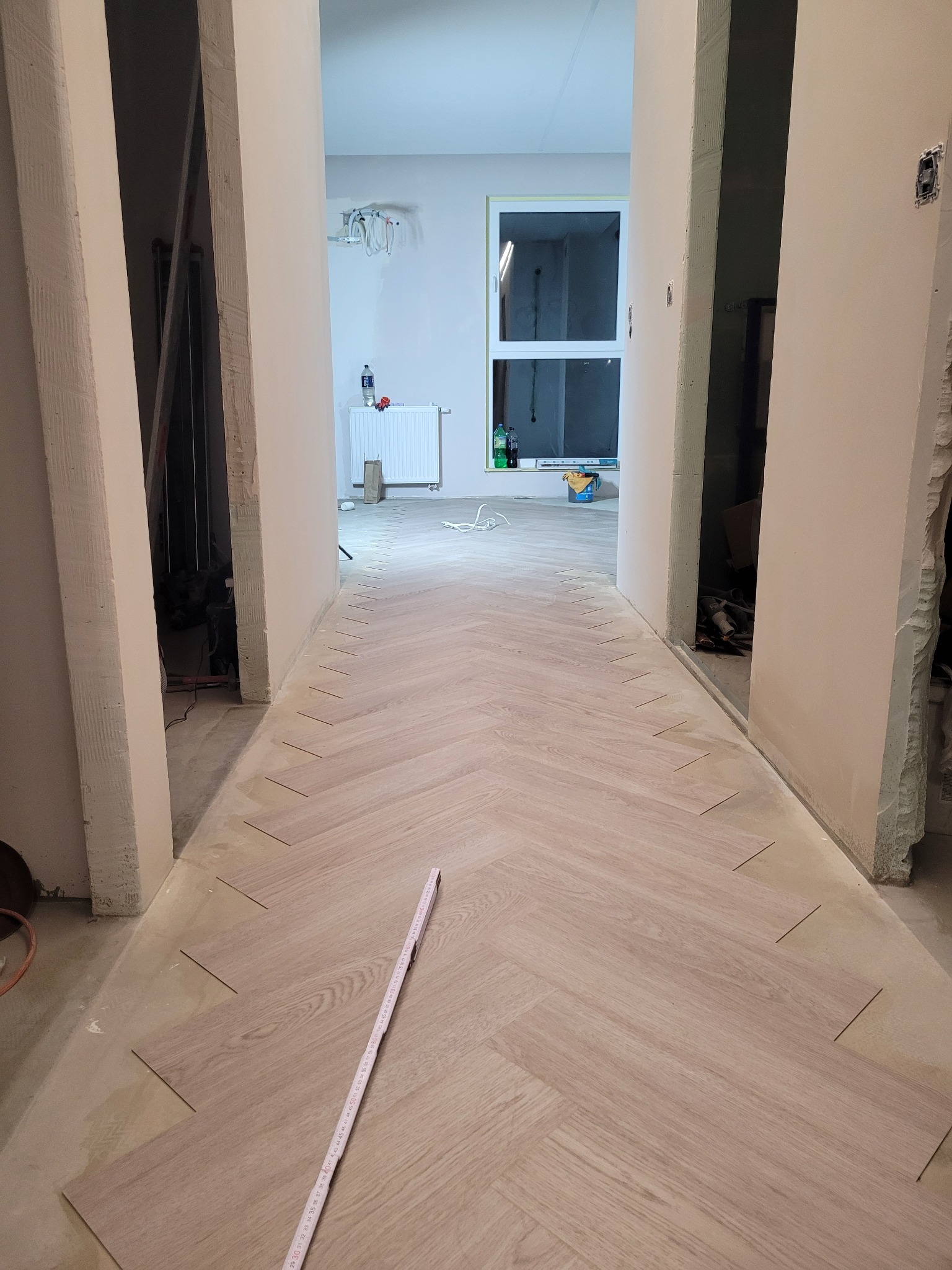 Układanie parkietu w jodełkę w korytarzu, widoczne fragmenty surowych ścian i ościeżnic, w tle okno i grzejnik, na podłodze leży miarka.