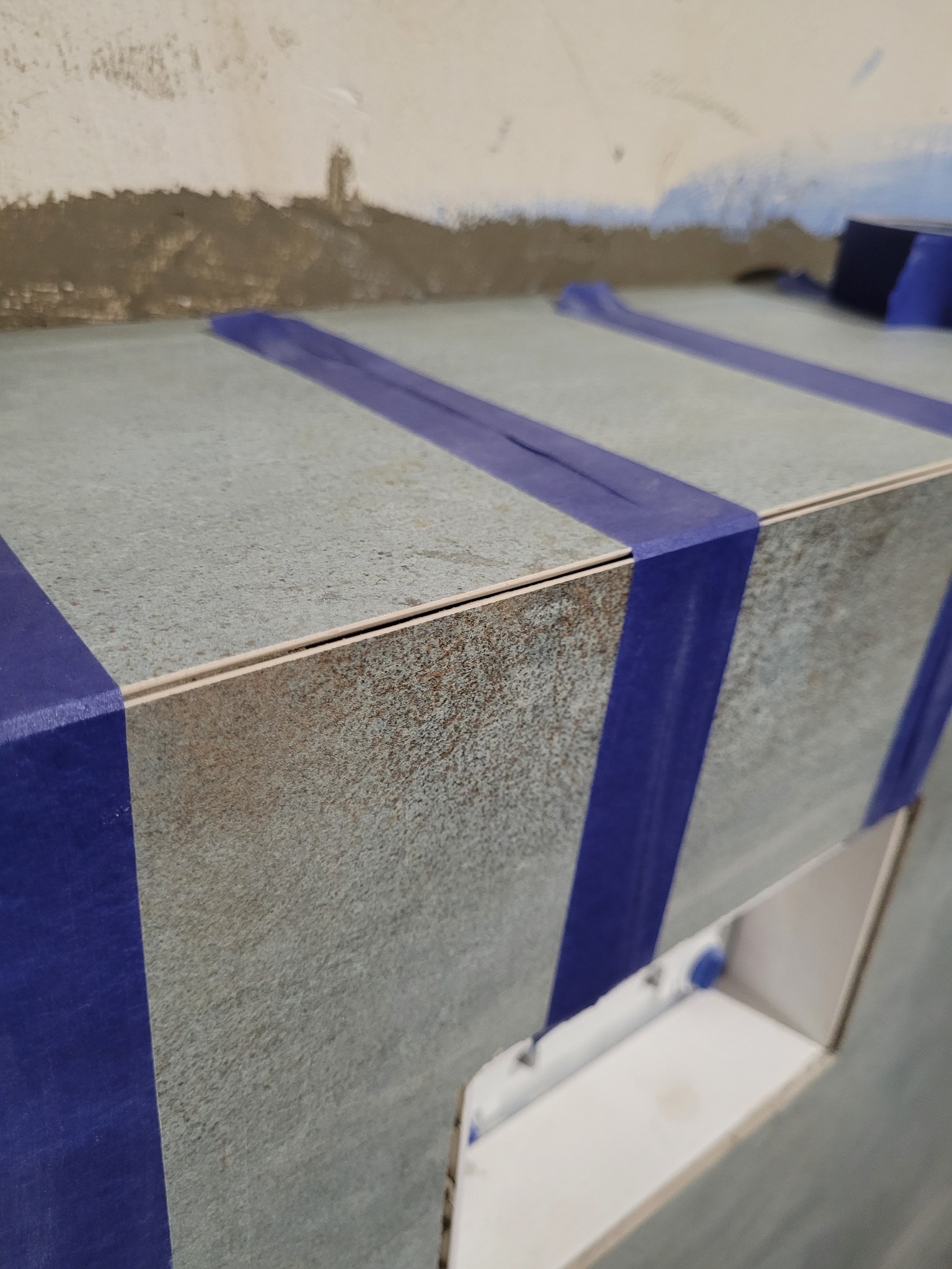 Montaż płytek ceramicznych na zabudowie w łazience, zabezpieczonych niebieską taśmą malarską, widoczne niedoskonałości na ścianie w tle.