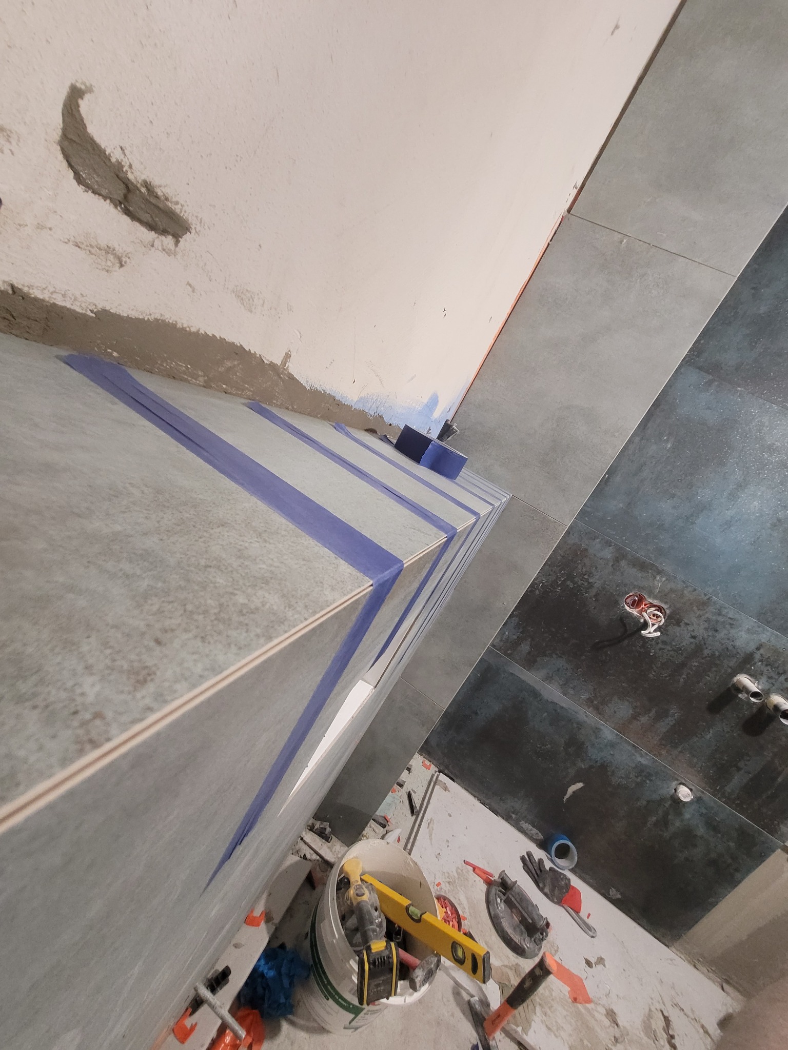 Świeżo ułożone, szare płytki ścienne zabezpieczone niebieską taśmą malarską, widoczne narzędzia pracy i poziomica w wiadrze, fragment surowej ściany z pozostałościami zaprawy.