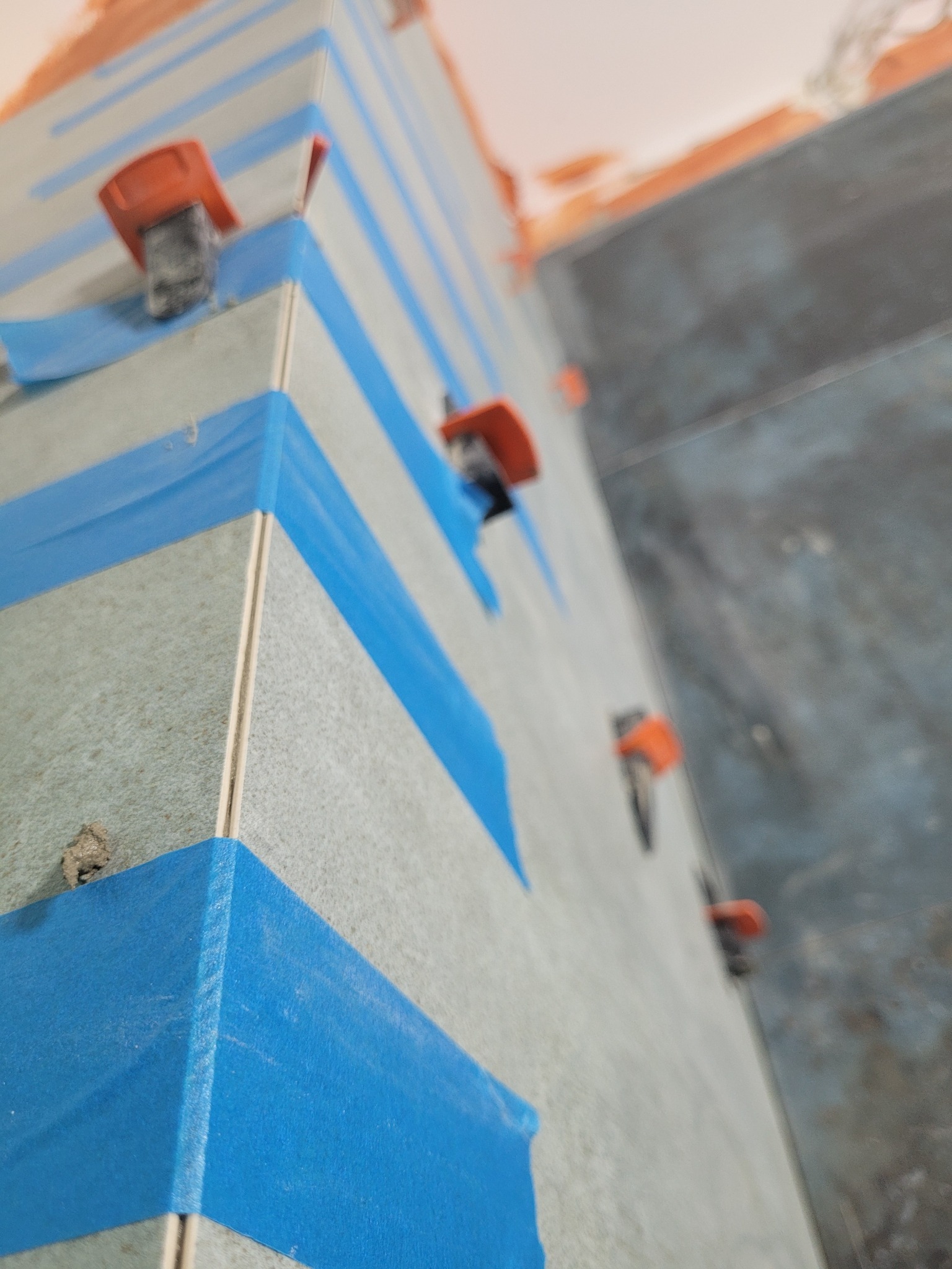 Fragment ściany w trakcie układania płytek, zabezpieczony niebieską taśmą malarską i poziomowany pomarańczowymi klipsami dystansowymi.