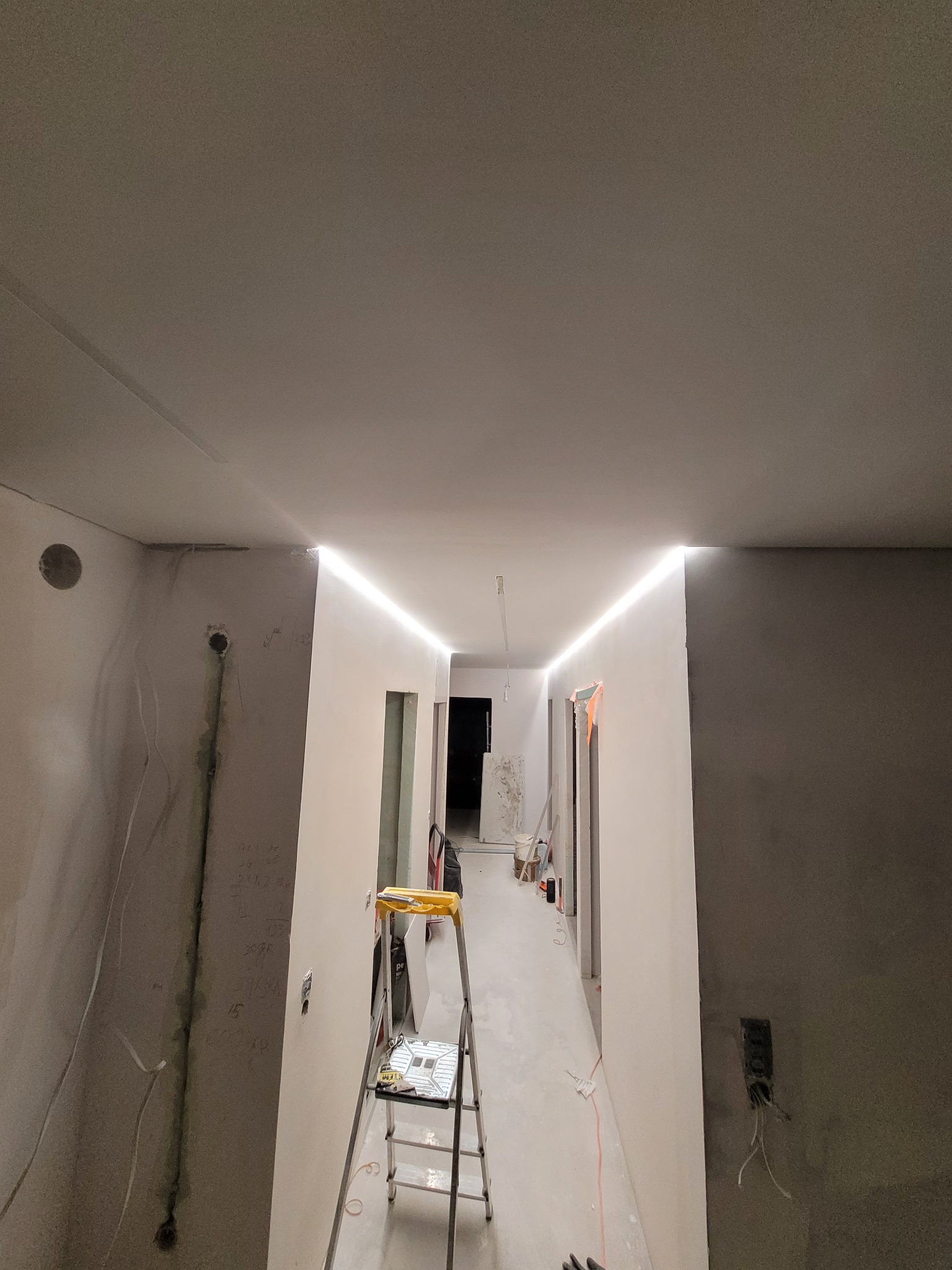Korytarz w trakcie prac wykończeniowych: widoczne surowe ściany, częściowo pomalowane, z instalacją elektryczną, schodki malarskie i oświetlenie LED wzdłuż sufitu.