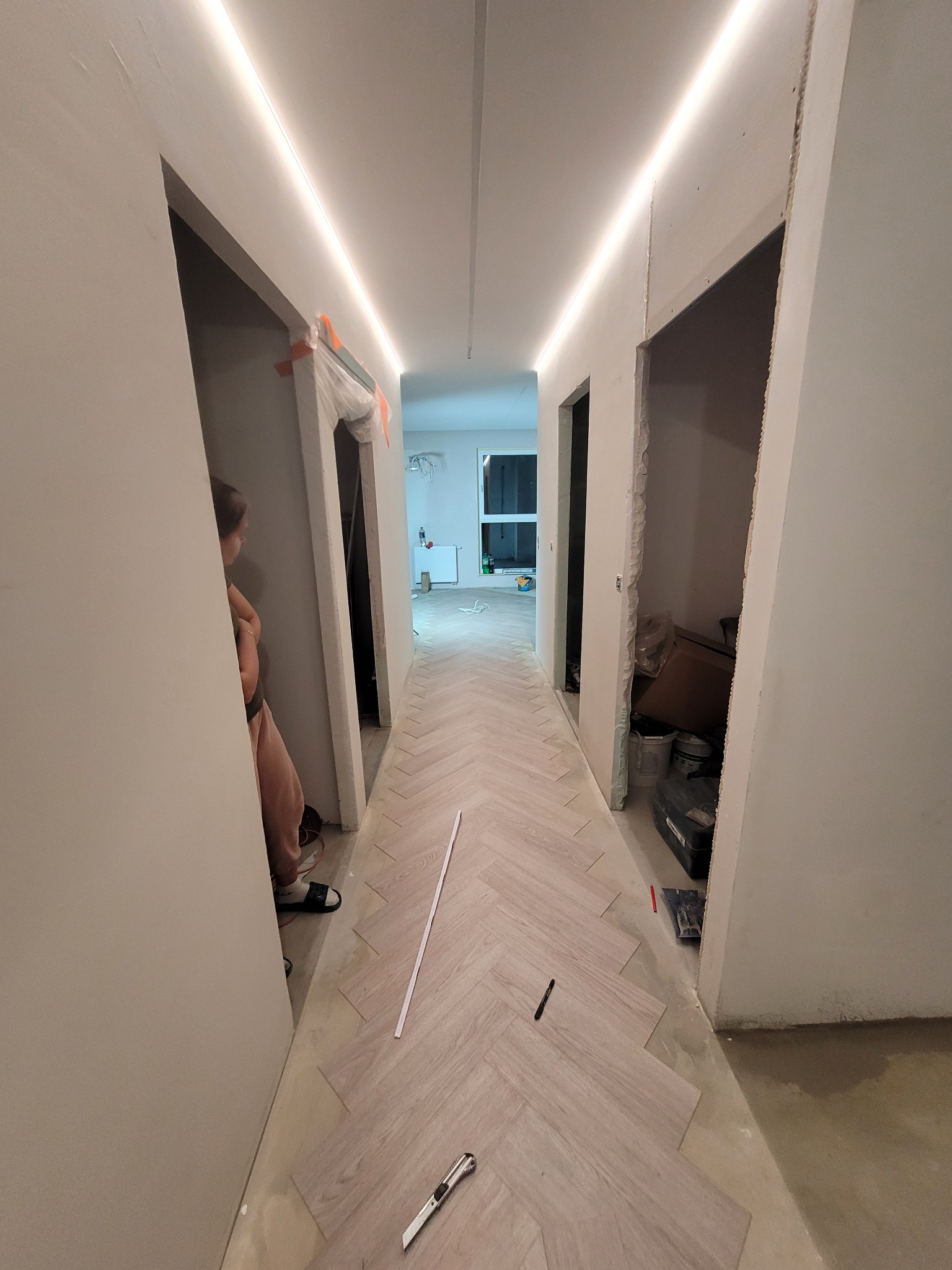 Korytarz w trakcie prac wykończeniowych: ułożona podłoga w jodełkę, częściowo zamontowane oświetlenie LED podsufitowe, widoczne narzędzia i materiały budowlane, ściany w trakcie przygotowania.