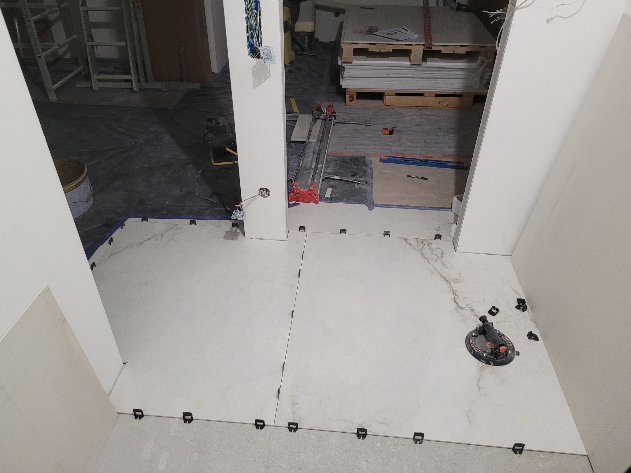 Układanie białych płytek podłogowych z marmurkowym wzorem, widoczne przyssawka do płytek i klipsy poziomujące spoiny, w tle narzędzia i płyty gipsowe.