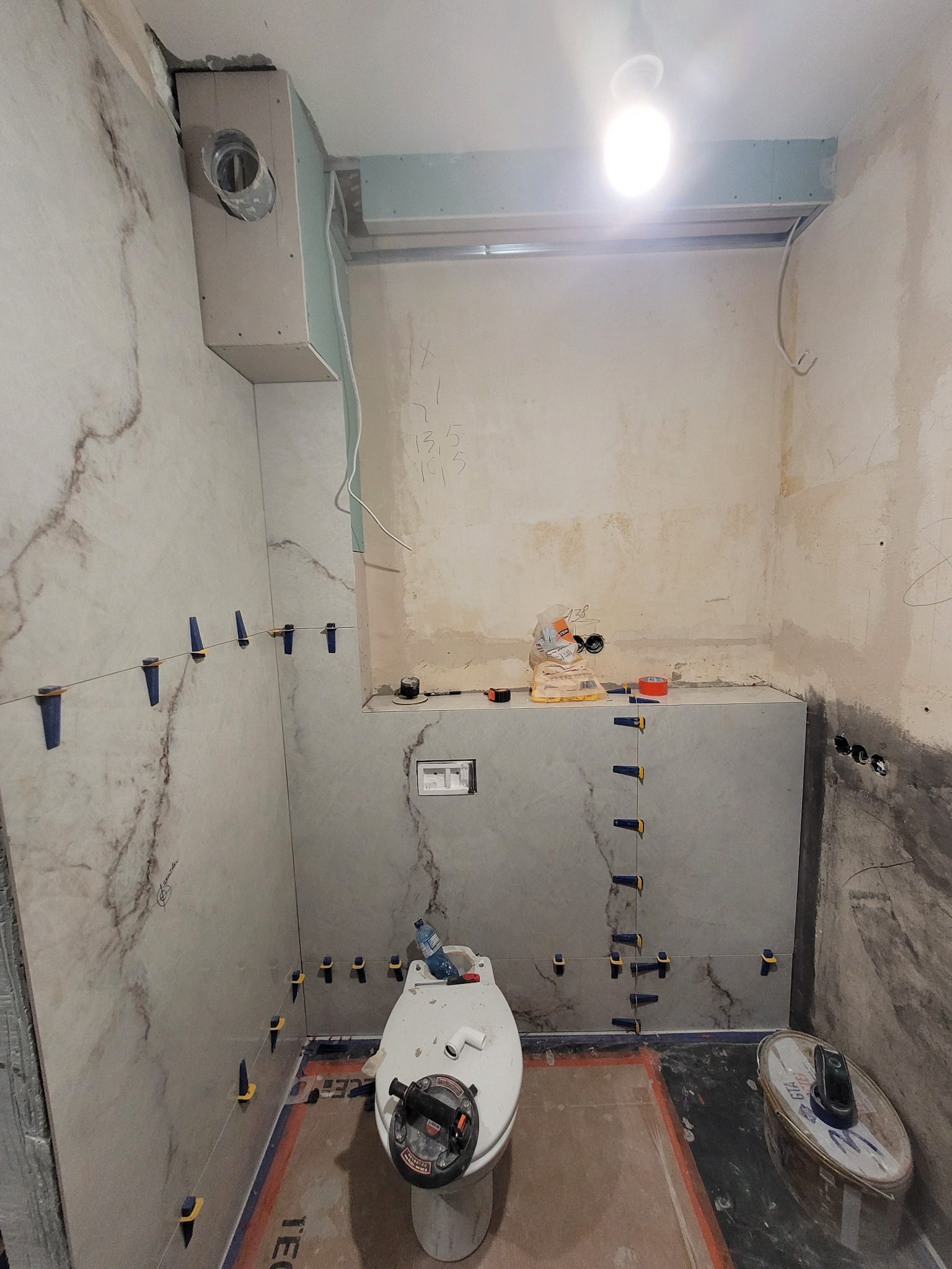 Remont łazienki: układanie płytek imitujących marmur na ścianach, z użyciem poziomowania i krzyżyków dystansowych, widoczna toaleta i narzędzia glazurnicze.