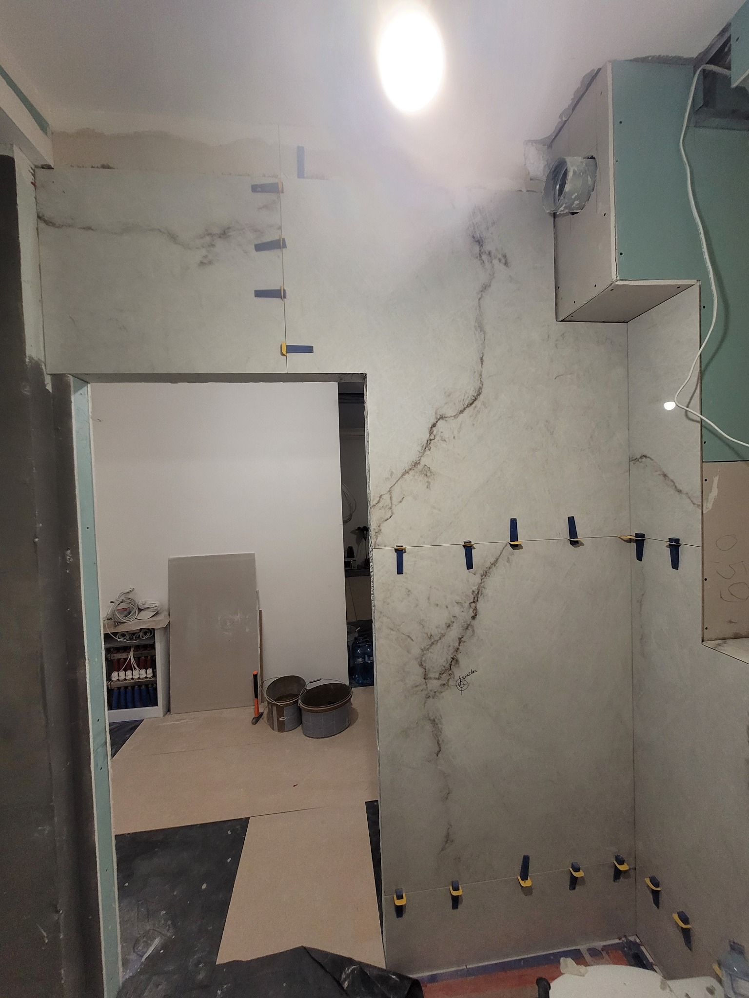 Ściana w trakcie układania płytek imitujących marmur, z użyciem klipsów poziomujących, widoczne płyty kartonowo-gipsowe i przygotowane podłoże w remontowanej łazience.