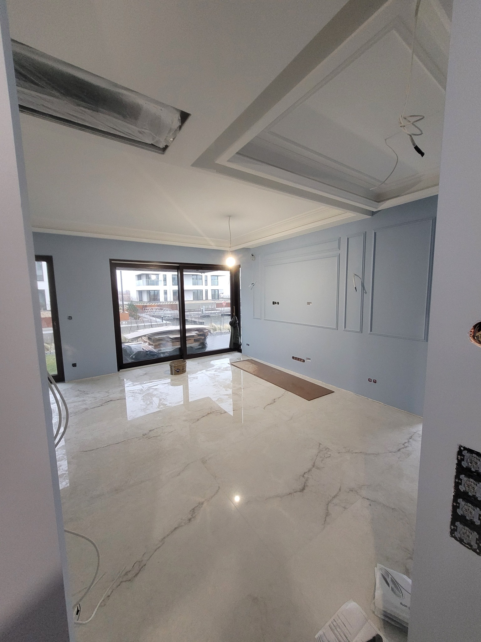 Jasne wnętrze w trakcie wykańczania, widoczne marmurowe płytki na podłodze, niebieskie ściany z dekoracyjnymi panelami, okno przesuwne z widokiem na budynek, zabezpieczone elementy sufitu folią.