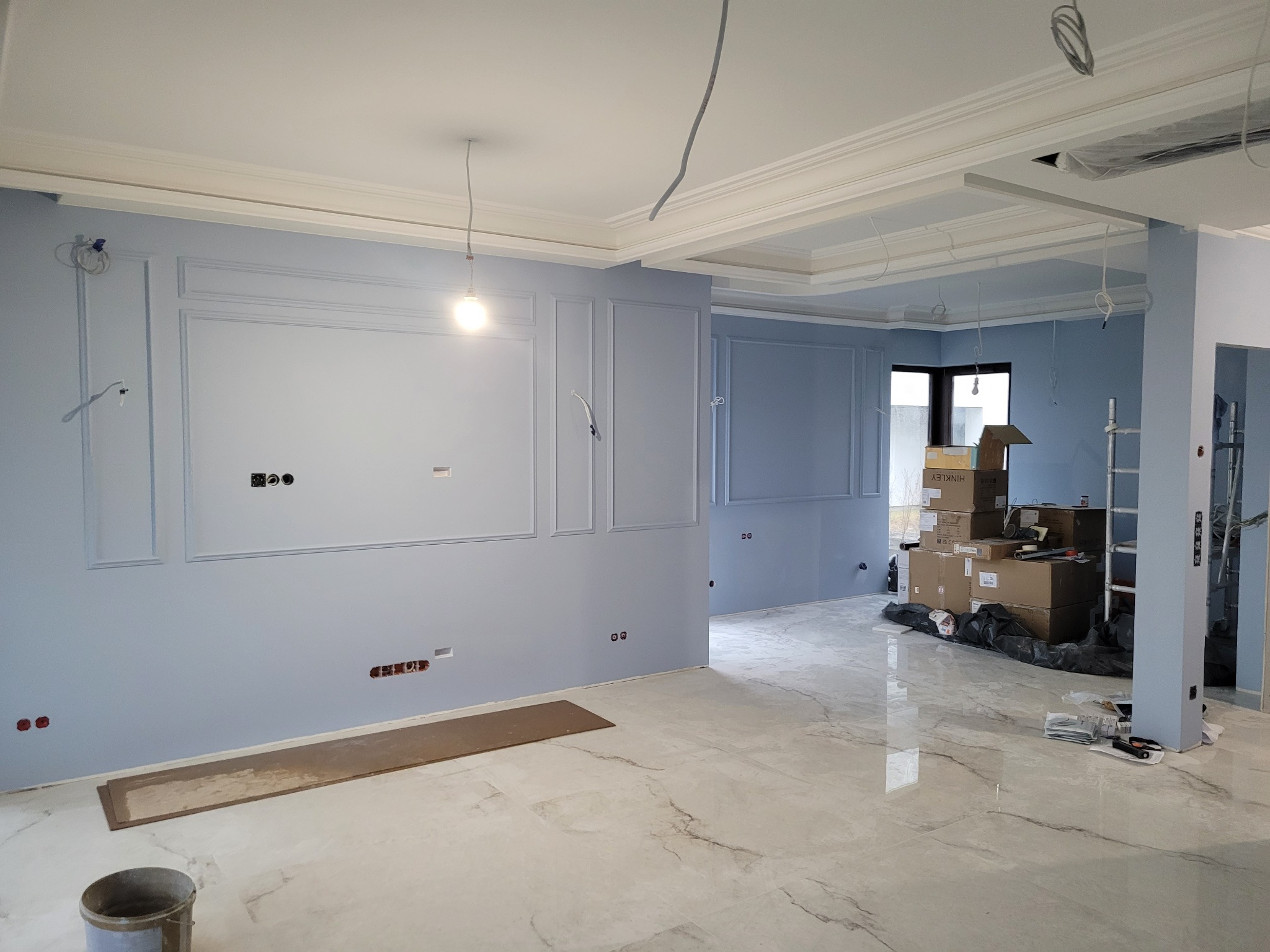 Wnętrze w trakcie prac wykończeniowych: ściany w kolorze gołębiej szarości z dekoracyjnymi listwami, marmurowe płytki na podłodze, widoczne puszki elektryczne i wystające przewody, kartony...
