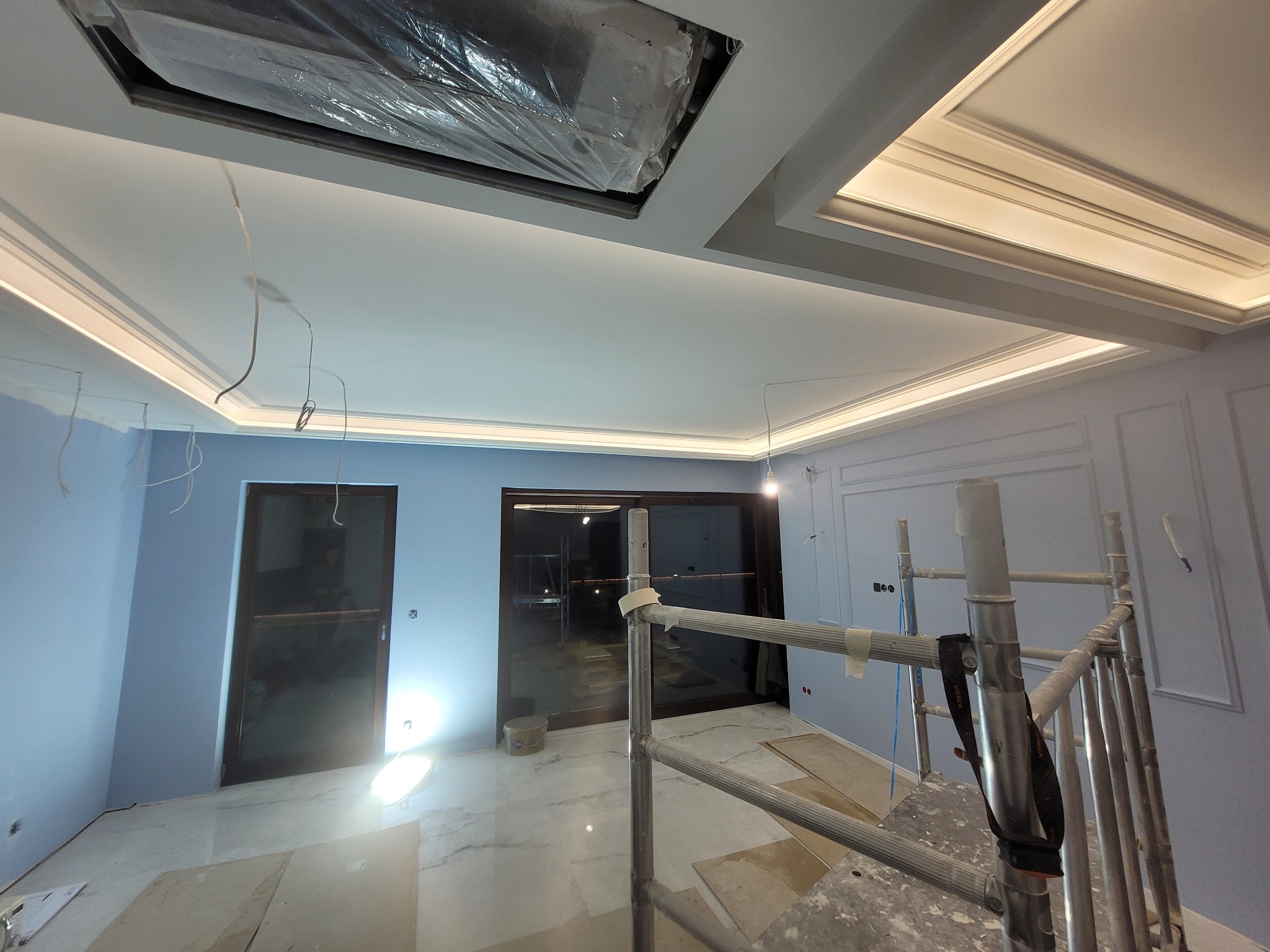 Wnętrze w trakcie prac wykończeniowych: pomalowane na niebiesko ściany z dekoracyjną sztukaterią, marmurowe płytki na podłodze, aluminiowe rusztowanie, podwieszany sufit z oświetleniem LED...