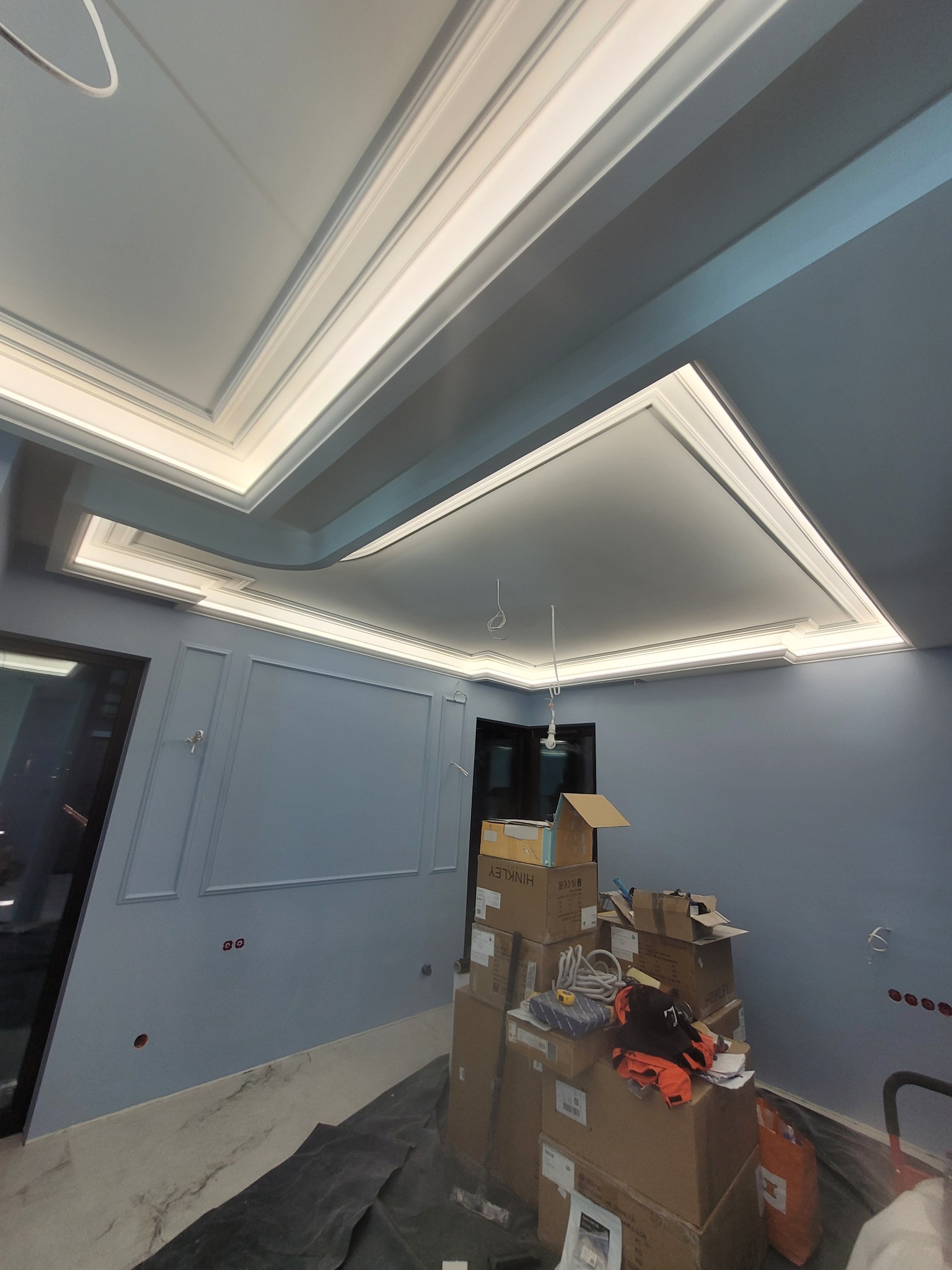 Wykończony sufit z oświetleniem LED w niebieskim pokoju, widoczne kartony i narzędzia budowlane świadczące o trwających pracach wykończeniowych.