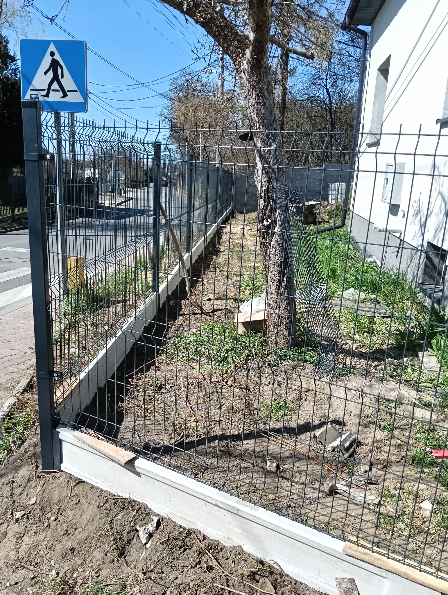 Montaż ogrodzenia panelowego z podmurówką betonową, widoczny znak przejścia dla pieszych i trawa rosnąca wzdłuż linii ogrodzenia.