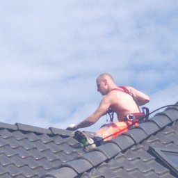 Fornalpol - Pracownik bez koszulki w uprzęży zabezpieczającej, wykonuje prace na ciemnym dachu z dachówki ceramicznej, widok z dołu na tle jasnego nieba.