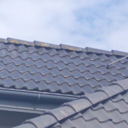 Fornalpol - Ciemna dachówka ceramiczna z widocznymi śladami mchu i zabrudzeń, częściowo przysłonięta przez rynnę, na tle błękitnego nieba.