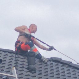 Fornalpol - Mężczyzna bez koszulki w uprzęży, z tatuażem na ramieniu, czyści dach z szarej dachówki za pomocą lancy ciśnieniowej; widoczna drabina oparta o dach.