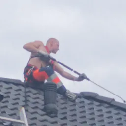 Mężczyzna bez koszulki w uprzęży, z tatuażem na ramieniu, czyści dach z szarej dachówki za pomocą lancy ciśnieniowej; widoczna drabina oparta o dach.